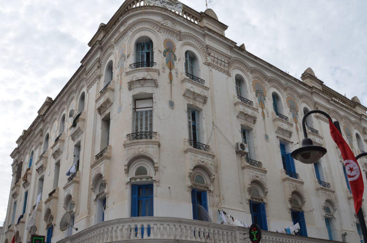 A photograph of a French-colonial style building in Tunisia. The building has arched windows, blue shudders, and a sign for a bar on the lower floor. A Tunisian flag is hanging on a lightpole in front og the building.