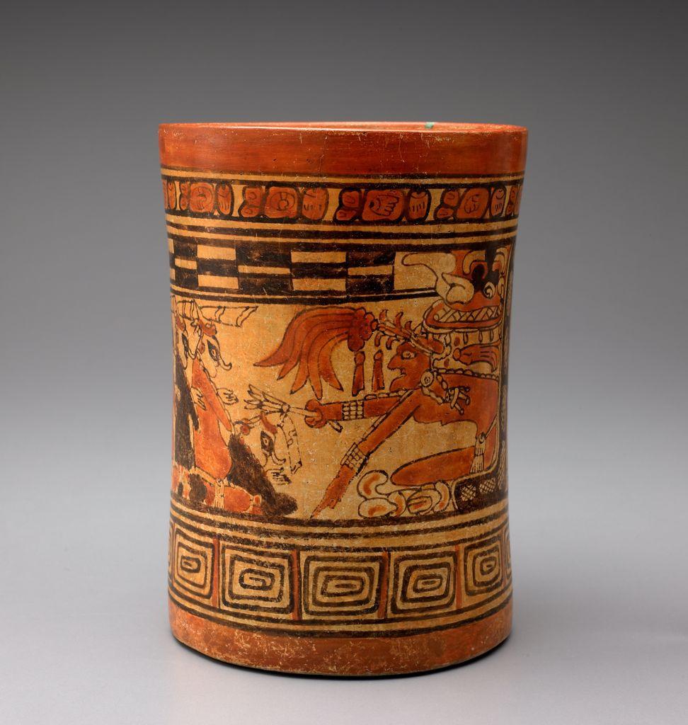 A ceramic vase depicting a humanoid figure pointing a reed-like object at a group of stylized humans, framed by repeating geometric motifs.
