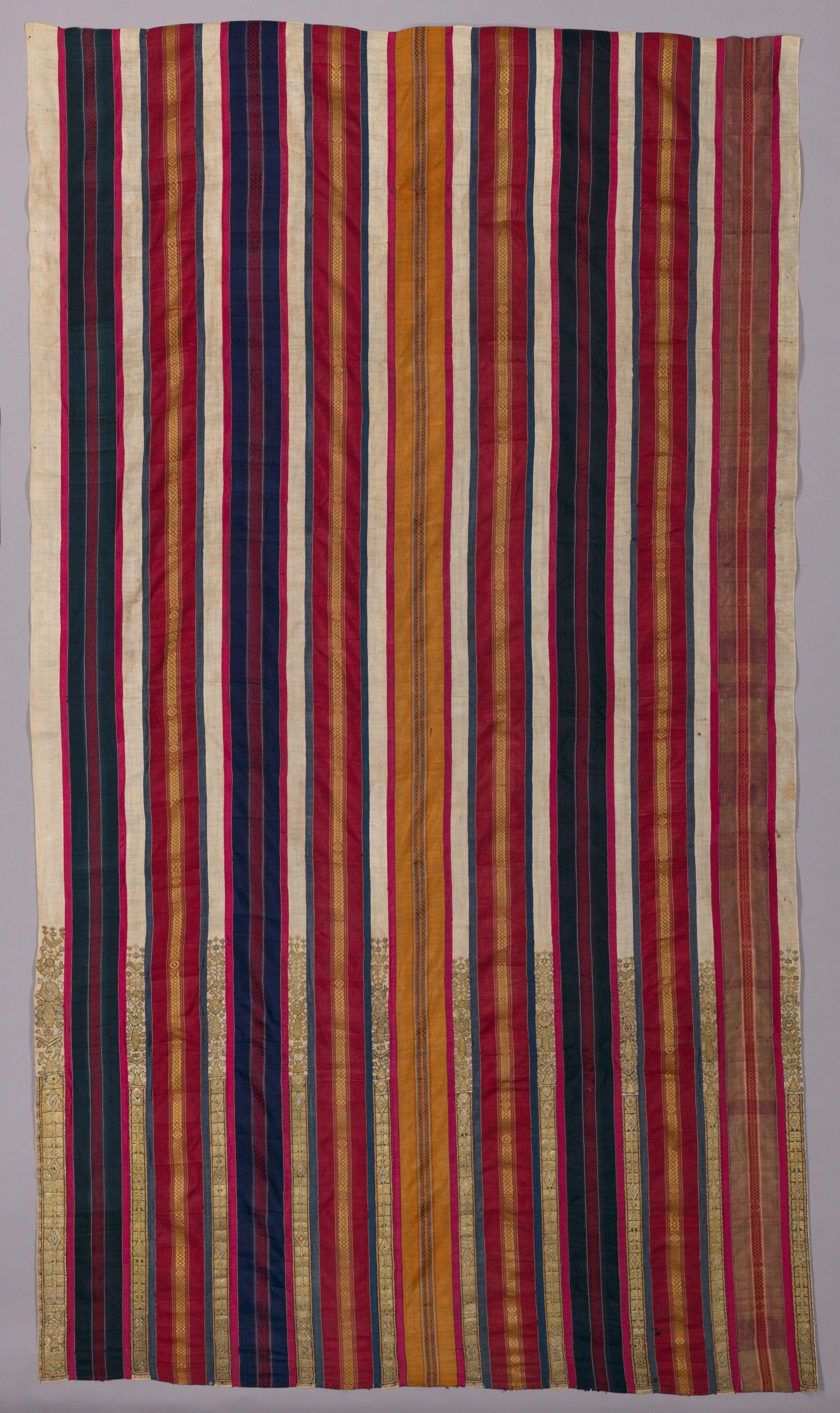 Photograph of a long, woven textile. There are narrow, alternative stripes of color. The colors include red, navy blue, mustard, tan, and black. Some of the bands have golden geometric and floral designs within them.